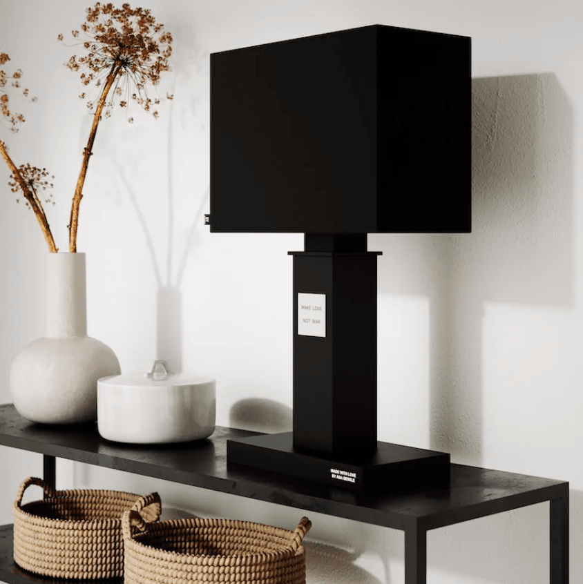 In Love 56cm Negro lámpara de mesa De By Rydéns - La amplia pantalla de tela negra contrasta armoniosamente con la estrecha base de la lámpara, que está decorada con una placa de metal con el texto “Haz el amor, no la guerra”.