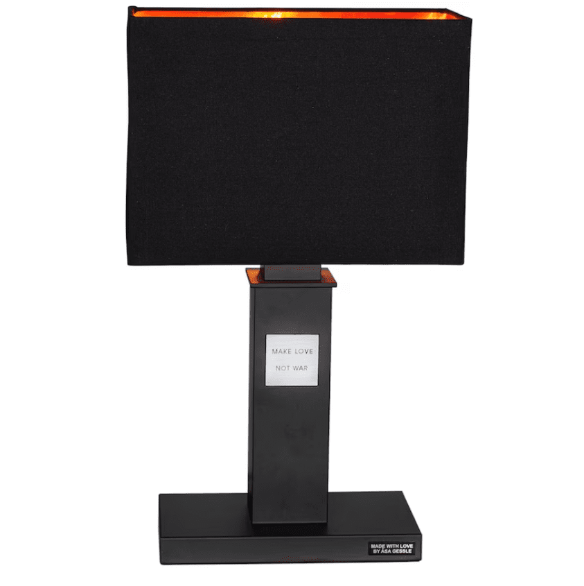 In Love 56cm Negro lámpara de mesa De By Rydéns - La amplia pantalla de tela negra contrasta armoniosamente con la estrecha base de la lámpara, que está decorada con una placa de metal con el texto “Haz el amor, no la guerra”.